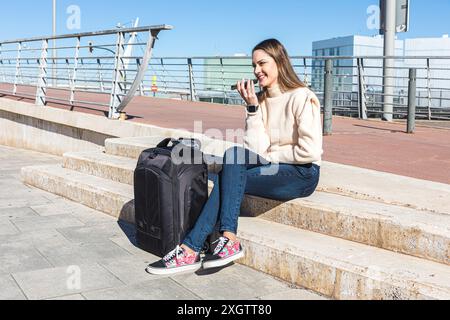 Eine junge Reisende sitzt auf den Stufen der Stadt, spricht glücklich mit ihrem Smartphone und blickt vor einer malerischen Kulisse der Stadt weg Stockfoto