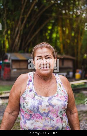 Eine ältere kolumbianische Frau, die ein Tanktop mit Blumenmuster trägt, steht selbstbewusst in einer ländlichen Landschaft. Üppiges Grün bildet eine einladende Kulisse Stockfoto