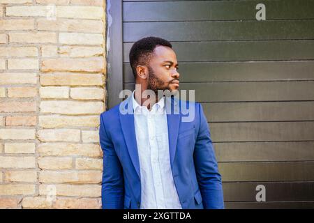 Ein Porträt eines besinnlichen jungen afroamerikanischen Mannes in einem stilvollen blauen Anzug, der vor einem strukturierten Backstein- und Metallhintergrund blickt. Stockfoto