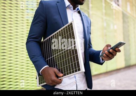 Ein nicht erkennbarer schwarzer Geschäftsmann, der ein Solarmodul und ein Smartphone hält und sich intensiv auf sein Gerät konzentriert. Der Mann ist stilvoll in Blau gekleidet Stockfoto