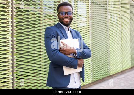 Ein lächelnder afroamerikanischer Geschäftsmann in einem stilvollen blauen Anzug, der selbstbewusst einen Laptop hält, während er in die Kamera schaut. Stockfoto