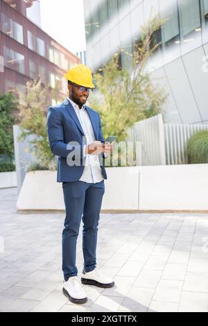 Ein afroamerikanischer Geschäftsmann in blauem Anzug und gelbem Schutzhelm steht draußen und schaut auf sein Smartphone. Die Szene hat eine moderne urbane Kulisse. Stockfoto