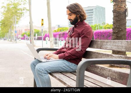 Ein bärtiger Mann in lässiger Kleidung benutzt einen Laptop, während er in Poblenou, Barcelona, auf einer Bank sitzt und Lifestyle und Arbeit mit einer sportlichen Stadtkulisse verbindet Stockfoto