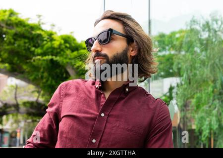 Ein modischer junger Mann in einem legeren kastanienbraunen Hemd und einer schwarzen Sonnenbrille steht draußen in Poblenou und verkörpert eine entspannte Lifestyle-Atmosphäre mit einem Hauch von s Stockfoto