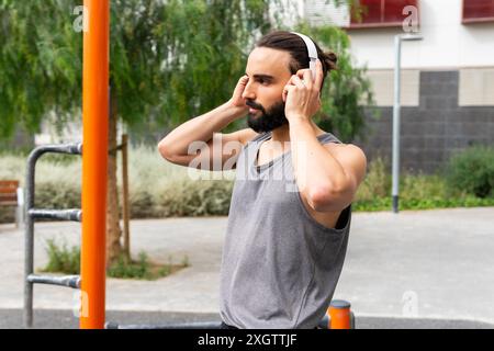 Ein fokussierter Mann bereitet sich auf seine Trainingsroutine vor, indem er seine kabellosen Kopfhörer in einem Fitnessbereich im Freien in Poblenou, Barcelona, anpasst und seine verbessert Stockfoto