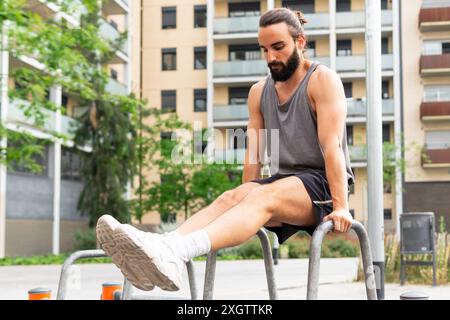 Ein fokussierter junger Mann mit Bart wird in einem Park in Poblenou, umgeben von modernen Wohngebäuden T, bei Beinübungen an parallelen Bars beobachtet Stockfoto