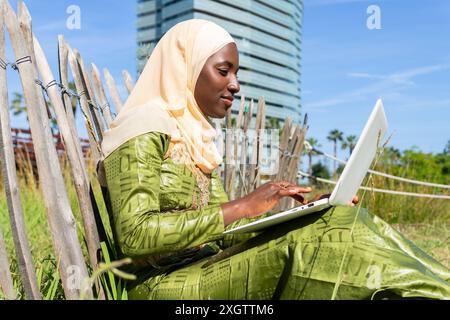 Eine junge muslimische Frau in einem Hijab arbeitet mit einem Laptop, während sie draußen in der Nähe eines hölzernen Zauns unter klarem Himmel sitzt Stockfoto