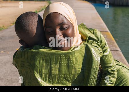 Ein herzerwärmender Moment von einer Mutter, die einen Hijab trägt, und ihrem jungen Sohn, der sich an einer ruhigen Uferpromenade in einem sonnendurchfluteten Park umarmt, ihr Ex Stockfoto