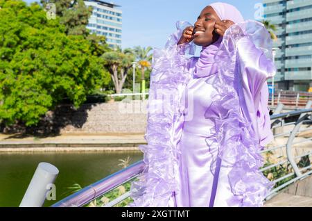 Eine lächelnde Frau in einem üppig violetten Hijab und Kleid steht fröhlich im Park, ihr Outfit fängt elegant das Sonnenlicht ein und reflektiert vib Stockfoto