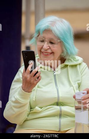 Eine fröhliche ältere Frau mit blauem Haar benutzt ihr Smartphone und genießt dabei Entspannung im Freien. Stockfoto