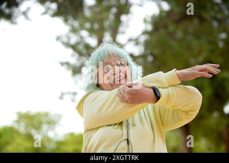 Von unten streckt eine fröhliche ältere Frau mit blauem Haar ihre Arme, während sie draußen trainiert, lächelt und wegblickt. Stockfoto
