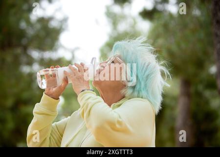 Seitenansicht einer älteren Frau mit blauen Haaren, die beim Sport im Freien aus einer Wasserflasche trinkt. Sie schaut weg. Stockfoto
