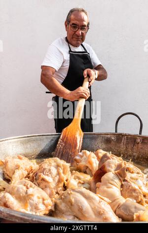 Ein reifer mexikanischer Mann, der Gläser und Schürze trägt, rührt eine große Pfanne kochender Hühner um und schaut auf das Essen herunter. Stockfoto