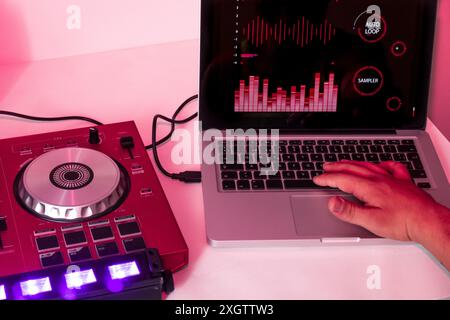 Nahaufnahme einer Hand, die Audioeinstellungen auf einem digitalen DJ-Mixer, der mit einem Laptop verbunden ist, abstimmt und Klangwellen unter rosa Beleuchtung anzeigt Stockfoto