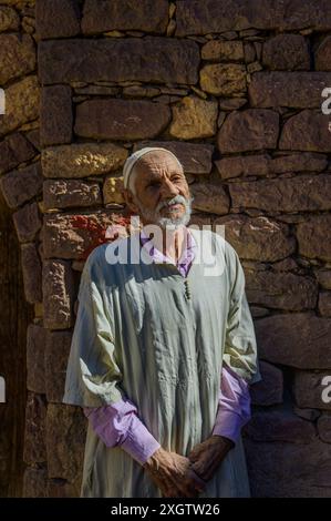 Im warmen Licht Marokkos steht ein älterer Mann in traditioneller Kleidung vor einer alten Steinmauer, die den zeitlosen Geist und Kultu verkörpert Stockfoto