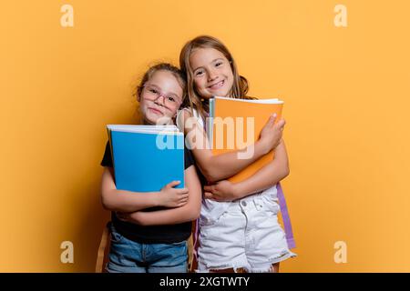 Zwei glückliche Mädchen, die sich umarmen, während sie bunte Bücher in einem Studio mit einem glatten gelben Hintergrund halten. Beide schauen in die Kamera und lächeln. Stockfoto