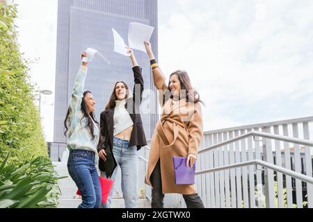 Drei junge Studentinnen feiern fröhlich mit Papieren in der Hand, draußen in der Nähe moderner städtischer Gebäude, die Glück und akademischen Erfolg darstellen Stockfoto