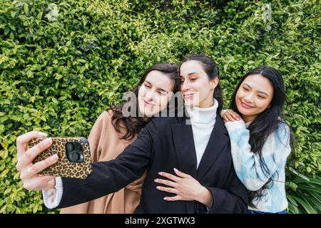 Drei fröhliche Schülerinnen machen gemeinsam ein Selfie vor einer üppigen grünen Hecke und zeigen ihre enge Freundschaft und ihr Glück Stockfoto