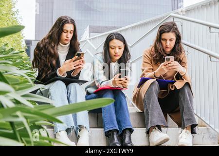 Drei junge Freundinnen sitzen eng auf einer Außentreppe, jede tief in ihr Smartphone verwickelt, umgeben von Stadtgebäuden Stockfoto