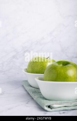 Leuchtend grüne Äpfel in eleganten weißen Schüsseln auf einer weichen grünen Serviette vor einem sauberen Marmorhintergrund. Diese Komposition hebt wh hervor Stockfoto