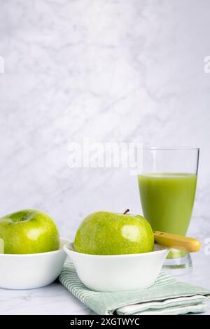 Frische grüne Äpfel in einer weißen Schüssel neben einem Glas nahrhaften Gurken- und Apfelsaft, elegant auf einer Marmoroberfläche platziert. Die Szene ist komplex Stockfoto