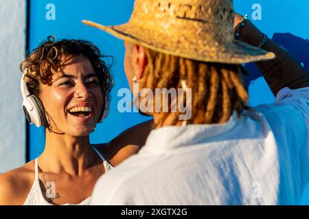 Eine fröhliche junge Frau mit lockigen Haaren und Kopfhörern führt ein lebhaftes Gespräch mit einem Mann, der einen Strohhut und Dreadlocks trägt. Beide sind lächelnd Stockfoto
