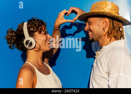 Ein kaukasischer Mann mit Dreadlocks und eine Frau mit lockigem Haar bilden mit ihren Händen vor einem leuchtend blauen Hintergrund eine Herzform. Beide lächeln und Stockfoto