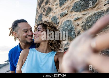 Ein Mann mit Dreadlocks und eine Frau mit lockigen Haaren, beide Kaukasier, lächeln und treten warm an einer zerklüfteten Steinmauer ein. Die Frau schaut fröhlich weg als Th Stockfoto