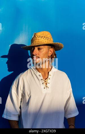 Ein junger kaukasischer Mann mit Dreadlocks, mit Strohhut und Schnürhemd, blickt nachdenklich vor einem leuchtend blauen Hintergrund weg. Stockfoto
