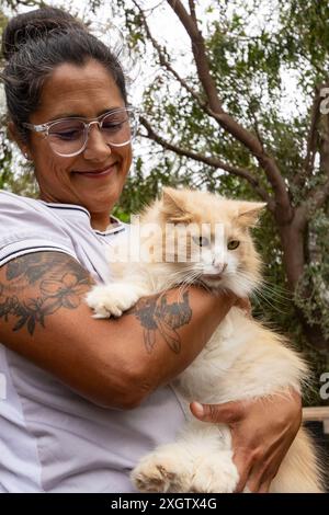 Eine Tierärztin in einem weißen Hemd mit sichtbaren Tätowierungen hält liebevoll eine flauschige weiße und beige Katze und lächelt, während sie hinunterblickt Stockfoto