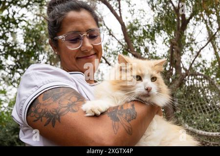 Von unten lächelt die Tierärztin sanft, während sie eine flauschige weiße und Ingwerkatze in ihren Armen hält. Beide sind im Freien, umgeben von Bäumen. Der Veter Stockfoto