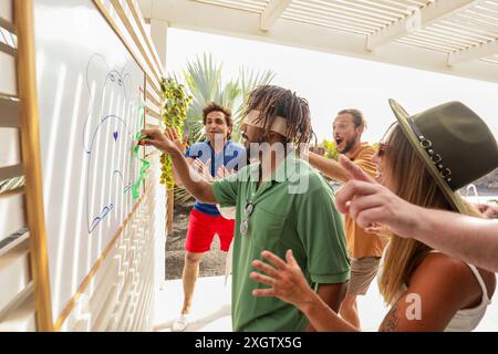 Eine Gruppe von Freunden genießt eine Sommerparty und spielt draußen ein Zeichenspiel mit verbundenen Augen. Aufregung und Lachen erfüllen die Luft, wenn sie jubeln und geben Stockfoto