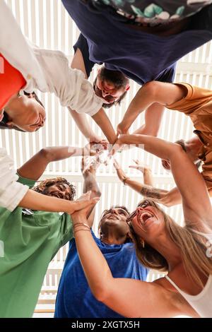 Von unten aus eine multiethnische Gruppe von Freunden, die lachen und mit ihren Händen ein Herz bilden, während sie in einem Kreis unter einem hellen, gestreiften Baldachin stehen. Stockfoto