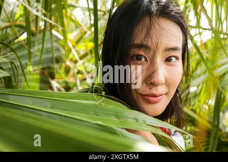 Nahaufnahme einer jungen asiatischen Frau, die in die Kamera schaut, während sie sanft durch leuchtend grüne Blätter blickt und einen verspielten, aber ruhigen Ausdruck zeigt. Stockfoto