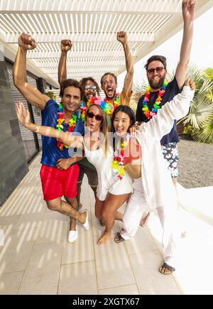 Eine lebendige und abwechslungsreiche Gruppe von sechs Freunden feiert den Sommer gemeinsam unter einer sonnigen Pergola. Sie sind in farbenfrohen Leis und lässigen Partyatti gekleidet Stockfoto