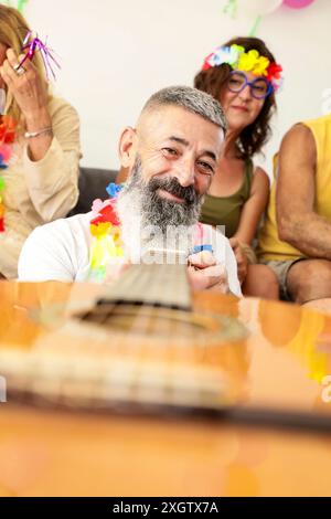 Eine fröhliche, reife Gruppe von Freunden genießt ein festliches Treffen, mit einem bärtigen Mann, der Gitarre als Mittelpunkt spielt, umgeben von Lachen und farbenfrohen p Stockfoto