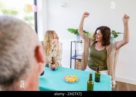 Drei Erwachsene werden bei einer zwanglosen Party im Haus gesehen. Eine Person tanzt fröhlich mit erhobenen Händen, während zwei andere dankbar lachen und sich unterhalten Stockfoto