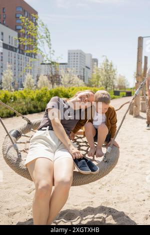 Eine Mutter und ihr Sohn haben einen freudigen Moment auf einer Schaukel im Park. Beide schauen nach unten, während sie zusammen lachen und den sonnigen Tag genießen. Städtische Gebäude Stockfoto