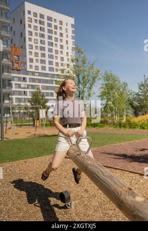 Junge Frau, die fröhlich auf einem hölzernen Tütler in einem Park spielt, umgeben von modernen Hochhäusern an einem sonnigen Tag. Fröhlich lachen, umarmen Stockfoto