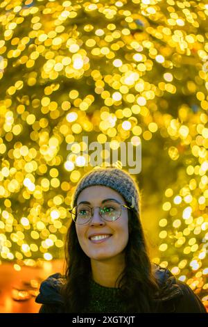 Ein Porträt einer fröhlichen jungen Frau mit Brille und Strickmütze, die warm lächelt und im Hintergrund leuchtende Weihnachtslichter glitzert Stockfoto