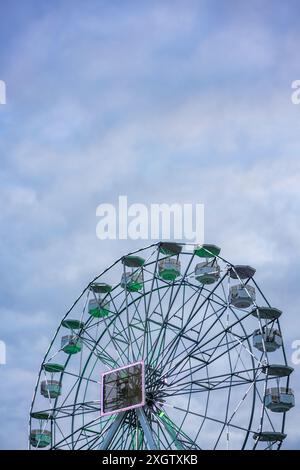 Ein Bild, das ein großes Riesenrad mit grünen und weißen Kabinen vor einem teilweise bewölkten blauen Himmel zeigt. Die Lichter des Gebäudes verleihen ein dezentes Leuchten. Stockfoto