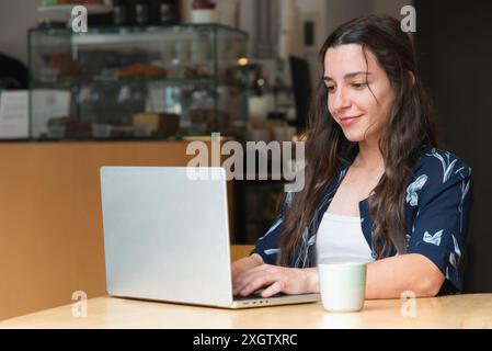 Eine junge Frau, die von der Kamera wegschaut, sitzt an einem Café-Tisch und arbeitet an ihrem Laptop neben einer Kaffeetasse, was auf eine produktive Kaffeepause hinweist. Stockfoto