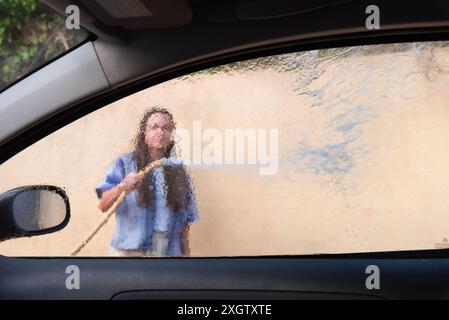 Blick aus dem Inneren eines Autos, der durch ein dampfendes Fenster hinausblickt und eine anonyme Frau zeigt, die das Auto wäscht. Sie scheint auf ihre Aufgabe konzentriert zu sein, nicht auf t zu schauen Stockfoto