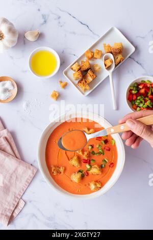 Blick von oben auf eine lebendige Schüssel mit traditioneller spanischer Tomaten-Gazpacho-Suppe, garniert mit Gemüse und Croutons. Ein Löffel schöpft die Suppe, hochlig Stockfoto