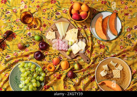 Ein lebhaftes, einladendes Picknick im Sommer mit einer Auswahl an Früchten, Käse und Wurstwaren auf einer gelben Tischdecke mit Blumenmuster. Stockfoto