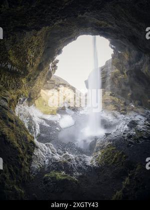 Ein fesselnder Blick auf einen Wasserfall, der von einem bogenförmigen Höhleneingang in Island herabstürzt, der mit Eiszapfen und Nebel verziert ist. Stockfoto