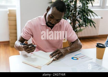 Ein afroamerikanischer Mann untersucht Dokumente in einem gut beleuchteten Coworking Space und zeigt organisierte Arbeiten mit Karten und einer Kaffeetasse in der Nähe. Stockfoto