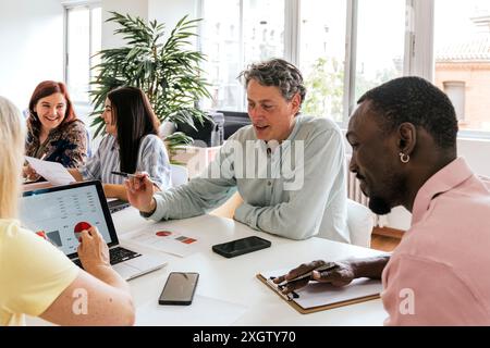 Eine multirassische Gruppe von Fachleuten arbeitet in einem sonnigen Coworking-Bereich zusammen. Das Team, bestehend aus Kaukasiern, Afrikanern und Hispanikern, wird von der Gruppe getrennt Stockfoto
