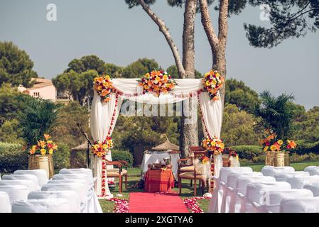 Elegante Hochzeitseinrichtung im Freien mit einem roten Teppich, der mit weißen Stühlen gesäumt ist, führt zu einem wunderschönen Blumenbogen, unter dem die Zeremonie stattfinden wird Stockfoto