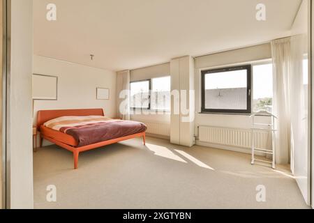 Modernes und luftiges Schlafzimmer mit einem großen Fenster mit Sonnenlicht in der Herentalsstraat, Amsterdam. Das Zimmer verfügt über ein orangefarbenes Bett, weiße Vorhänge und eine Meile Stockfoto